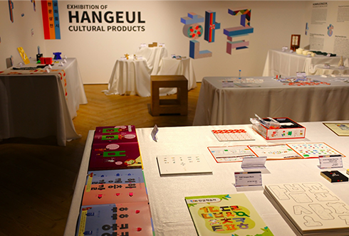 한글문화산업전 사진이다. 벽면에는 ‘Exhibition of Hangeul Cultural Products’가 적혀 있고, 여러 색으로 장식된 ‘한글’이라는 글자가 그 옆에 배치되어 있다. 흰색 테이블에는 한글이 적혀 있는 여러 작품이 전시되어 있다.