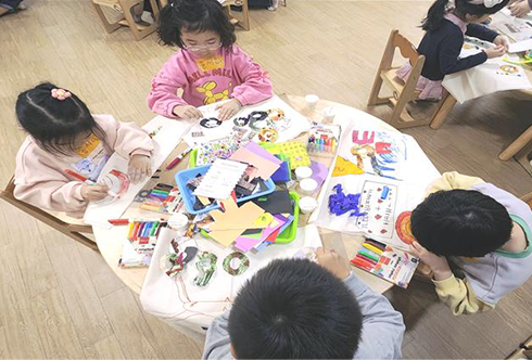 한글 기반 문화예술 융합 교육 시범 운영 현장 사진이다. 흰색 둥근 탁자에 네 명의 어린아이가 둘러앉아 색연필과 크레파스로 그림을 그리고 있다. 탁자 위에는 종이와 색연필, 사인펜 등 여러 미술용품이 가득 쌓여있다.