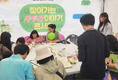 ‘찾아가는 사투리 이야기 콘서트’ 연계 한글문화 체험 행사 사진이다. 여러 사람이 큰 탁자에 둘러앉아 종이에 무언가를 그리거나 적고 있다. 행사 진행 요원 성인 두 명은 체험 행사에 참여한 사람들을 바라보며 안내하고 있다. 