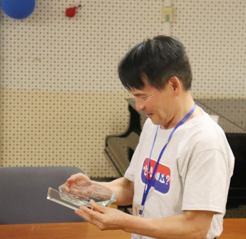 반갑습니다 사진. ‘안녕하세요?’가 적힌 흰색 반팔을 입고 있는 강병구 교수가 들고 있는 상패를 내려다보며 웃고 있다.