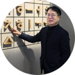 국립한글박물관 강정원 관장 사진이다. 안경을 쓰고 있는 강정원 관장이 팔짱을 끼고 부드럽게 미소 짓고 있다. 검은색 터틀넥과 짙은 남색 정장을 입었으며 오른쪽 손에 손목시계를 차고 있다. 그의 뒤에는 국립한글박물관 상설전시 작품 중 하나인 훈민정음해례본 원형 33장을 활용한 설치 조형물이 놓여 있다.