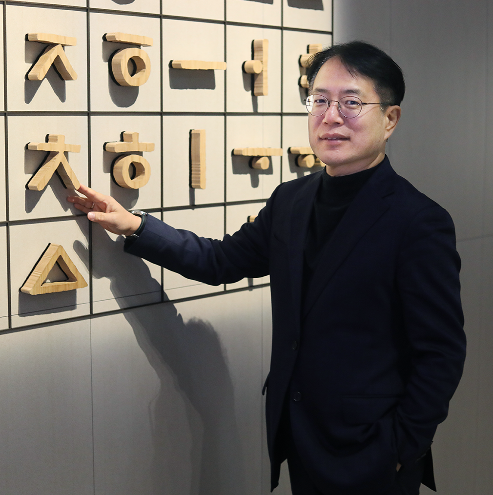 기획 기사 사진. 국립한글박물관 강정원 관장 사진이다. 안경을 쓰고 있는 강정원 관장이 팔짱을 끼고 부드럽게 미소 짓고 있다. 검은색 터틀넥과 짙은 남색 정장을 입었으며 오른쪽 손에 손목시계를 차고 있다. 그의 뒤에는 국립한글박물관 상설전시 작품 중 하나인 훈민정음해례본 원형 33장을 활용한 설치 조형물이 놓여 있다.