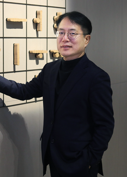 기획 기사 사진. 국립한글박물관 강정원 관장 사진이다. 안경을 쓰고 있는 강정원 관장이 팔짱을 끼고 부드럽게 미소 짓고 있다. 검은색 터틀넥과 짙은 남색 정장을 입었으며 오른쪽 손에 손목시계를 차고 있다. 그의 뒤에는 국립한글박물관 상설전시 작품 중 하나인 훈민정음해례본 원형 33장을 활용한 설치 조형물이 놓여 있다.