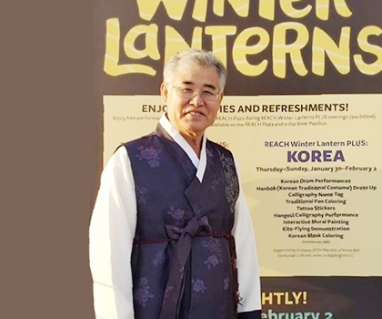 반갑습니다 사진. 진한 보라색 한복을 입고 웃고 있는 권명원 서예가 사진이다. 권명원 서예가는 무테안경을 쓰고 있으며 카메라를 바라보며 환하게 웃고 있다. 배경에는 영어가 쓰인 현수막이 있다.