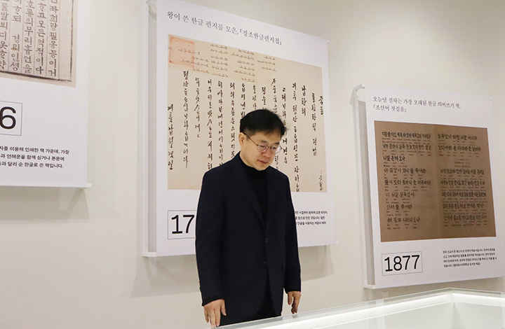 강정원 관장이 국립한글박물관 유리 진열대에 전시된 유물을 내려다보고 있다. 그의 뒤쪽에는 ‘왕이 쓴 한글 편지들 보존, 「정조한글편지첩」 등 한글 유물 관련 전시 패널이 걸려있다. 