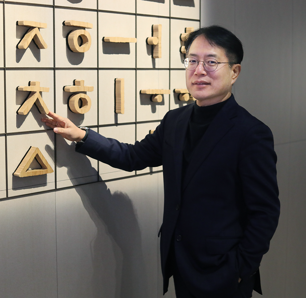 기획기사 사진. 국립한글박물관 강정원 관장 사진이다. 안경을 쓰고 있는 강정원 관장이 팔짱을 끼고 부드럽게 미소 짓고 있다. 검은색 터틀넥과 짙은 남색 정장을 입었으며 오른쪽 손에 손목시계를 차고 있다. 그의 뒤에는 국립한글박물관 상설전시 작품 중 하나인 훈민정음해례본 원형 33장을 활용한 설치 조형물이 놓여 있다.