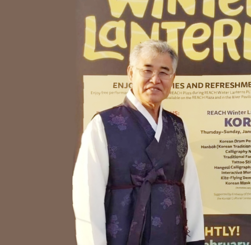반갑습니다 사진. 진한 보라색 한복을 입고 웃고 있는 권명원 서예가 사진이다. 권명원 서예가는 무테안경을 쓰고 있으며 카메라를 바라보며 환하게 웃고 있다. 배경에는 영어가 쓰인 현수막이 있다.