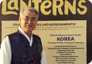 진한 보라색 한복을 입고 웃고 있는 권명원 서예가 사진이다. 권명원 서예가는 무테안경을 쓰고 있으며 카메라를 바라보며 환하게 웃고 있다. 배경에는 영어가 쓰인 현수막이 있다. 