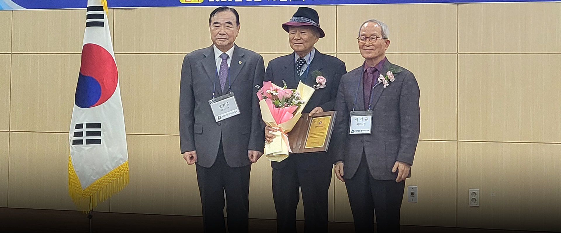 반갑습니다 사진. 양복을 입은 세 남성이 나란히 서 있다. 오른쪽 남성은 짙은 회색 양복을 입고 있고, 이름표를 목에 걸고 있다. 가운데 남성은 (사)한국시조협회 김달호 세계화위원장으로, 중절모를 쓰고 있다. 한 손에는 꽃다발, 다른 한 손에는 표창장을 들고 서 있다. 왼쪽 남성 역시 정장 차림이며, 그의 옷에는 핀으로 고정된 작은 꽃장식이 있다.