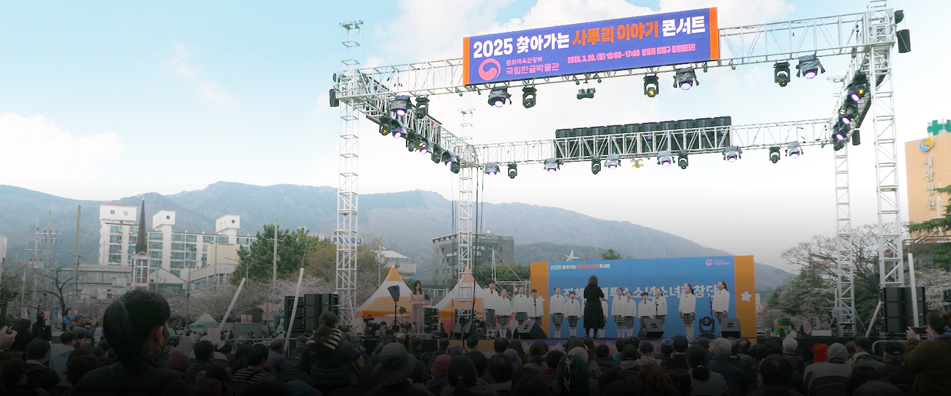 ‘2025 찾아가는 사투리 이야기 콘서트’ 행사 사진이다. 무대 위쪽에는 가로로 긴 현수막이 걸려 있으며, ‘2025 찾아가는 사투리 이야기 콘서트’라는 문구가 적혀있다. 무대에는 흰색 상의와 체크무늬 하의를 맞춰 입은 진해안젤루스 소년소녀 합창단원들이 일렬로 서 있다. 관객석에는 다양한 연령대의 사람들이 앉아 있다. 왼쪽 하단에는 하늘색 박스 안에 ‘기획기사’ 문구가 적혀있다. 하단에는 기사의 제목 ‘진해 군항제와 함께한 봄맞이 현장 찾아가는 사투리 이야기 콘서트’가 적혀있다.