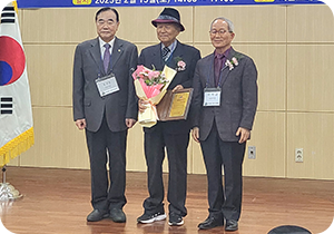 양복을 입은 세 남성이 나란히 서 있다. 오른쪽 남성은 짙은 회색 양복을 입고 있고, 이름표를 목에 걸고 있다. 가운데 남성은 (사)한국시조협회 김달호 세계화위원장으로, 중절모를 쓰고 있다. 한 손에는 꽃다발, 다른 한 손에는 표창장을 들고 서 있다. 왼쪽 남성 역시 정장 차림이며, 그의 옷에는 핀으로 고정된 작은 꽃장식이 있다.
