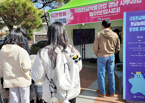 한글문화 체험 부스 현장 사진이다. 야외에 설치된 부스 위에 ‘국립한글박물관과 경남 사투리랑 놀아보입시더!’라고 적혀있다. 부스 앞에는 흰색 옷을 입은 학생들이 서있다.