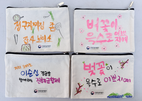 나만의 사투리 주머니 만들기 체험에 참여한 사람들 작품이다. 연한 베이지색 파우치에 각자 손 글씨를 적고 그림을 그려 꾸몄다. 왼쪽 상단에는 ‘정구지찌짐 잡사보이소’라고 적혀 있고, 오른쪽 상단에는 ‘버꽃이 윽수로 이쁘지예’라고 적혀있다. 왼쪽 하단에는 ‘자리 오이소 이순신 장군캄 함께하는 진해군항제’라고 쓰여있고, 오른쪽 하단에는 ‘벚꽃이 윽수로 이쁘지예’라고 적혀있다. 