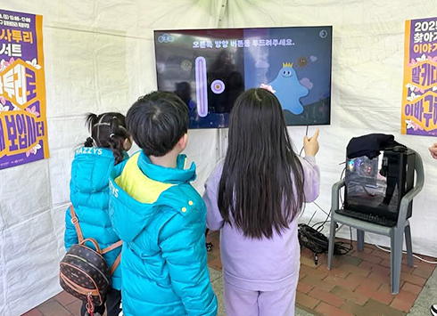 한글 자음과 모음ㆍ합자의 원리를 익히는 영상 게임 영상 체험 현장이다. 하늘색 패딩을 입고 있는 여자아이와 남자아이, 보라색 옷을 입고 있는 여자아이가 모니터를 바라보며 게임을 하고 있다. 그 옆에는 관계자로 보이는 검은색 패딩을 입은 남성이 아이들이 게임하는 걸 바라보고 있다.