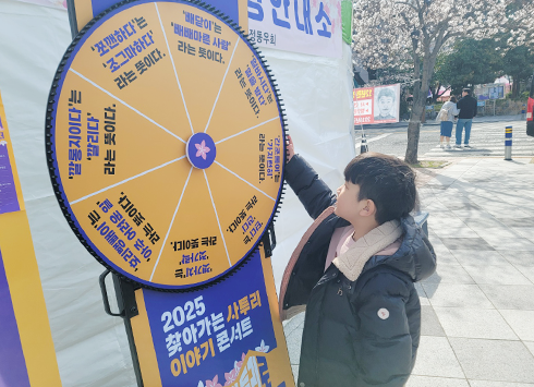 원판을 돌려 사투리 문제 맞히기 체험 현장이다. 검은색 패딩을 입은 남자아이가 주황색 원판 룰렛을 손으로 돌리고 있다.