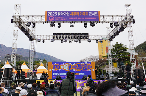 콘서트 시작 전, 객석을 가득 메운 관람객들 사진이다. 야외에 설치된 무대 위에는 행사 현수막이 걸려 있다. 무대 배경에는 시선을 사로잡는 독특한 입체 디자인으로 ‘말키다 사투리로 이바구해 보입시더’가 적혀있다. 관객석은 사람들로 인해 꽉 차 있다.