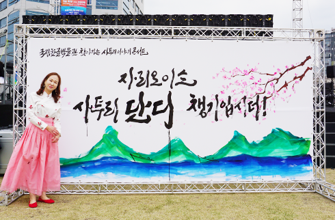 경남 사투리로 손 멋글씨 그린 정윤희 작가 사진이다. 대형 손글씨 배너 앞에서 포즈를 취하고 있는 정윤희 작가의 모습이 담겨있다. 배너에는 ‘자리 오이소 사투리로 단디 챙기입시더!’가 붓글씨로 크게 적혀있다. 배경에는 산과 벚꽃을 수채화로 표현한 그림이 함께 어우러져 있다. 정윤희 작가는 분홍색 치마와 흰 저고리를 입고 활짝 웃고 있다.