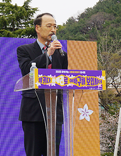 김정대 경남대 명예교수님 사진이다. 연단 앞에 서 있는 김정대 경남대 명예교수가 마이크를 들고 말하고 있다. 검은 정장에 붉은 계열의 넥타이를 착용했으며 안경을 쓰고 있다.