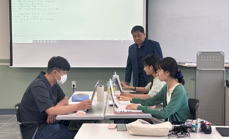 예비 교사들에게 점자 지도를 하고 있는 박중휘 교수 사진이다. 가운데 테이블에 세명의 학생이 앉아 노트북 화면을 들여다보고 있다. 앞쪽 스크린에는 점자로 작성된 문서가 띄워져 있다. 