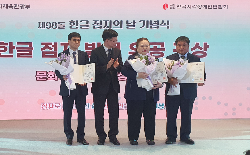 한글 점자 발전 유공자로 선정된 박중휘 교수 사진이다. 제98돌 한글 점자의 날 기념식에서 한글 점자 발전 유공자로 선정된 박중휘 교수 외 두 명이 상패와 꽃다발을 들고 서 있다.