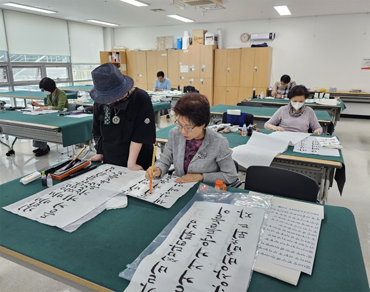 실내 공간에서 한글 서예 수업이 이루어지고 있는 사진이다. 밝은 조명이 켜진 교실 같은 실내에는 초록색 천이 덮인 테이블들이 줄지어 놓여 있다. 여러 명의 사람이 각자 자리에 앉아 한글 서예를 연습하고 있습니다. 사진 중앙에는 두 명의 여성이 함께 서예 작품을 보고 있다. 테이블에 앉아 있는 여성은 회색 정장을 입고 안경을 썼으며, 오른손에 붓을 들고 정성스럽게 붓글씨를 쓰고 있다. 왼쪽에는 검은 모자를 쓴 또 다른 여성이 옆에서 한글 서예를 지도하는 듯한 모습이다.