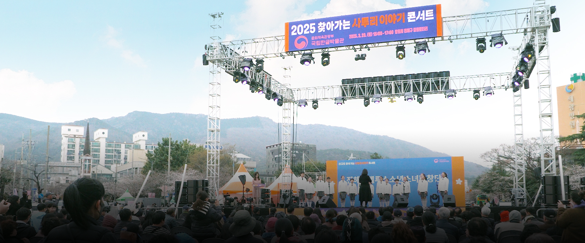 기획기사 사진. ‘2025 찾아가는 사투리 이야기 콘서트’ 행사 사진이다. 무대 위쪽에는 가로로 긴 현수막이 걸려 있으며, ‘2025 찾아가는 사투리 이야기 콘서트’라는 문구가 적혀있다. 무대에는 흰색 상의와 체크무늬 하의를 맞춰 입은 진해안젤루스 소년소녀 합창단원들이 일렬로 서 있다. 관객석에는 다양한 연령대의 사람들이 앉아 있다. 왼쪽 하단에는 하늘색 박스 안에 ‘기획기사’ 문구가 적혀있다. 하단에는 기사의 제목 ‘진해 군항제와 함께한 봄맞이 현장 찾아가는 사투리 이야기 콘서트’가 적혀있다.