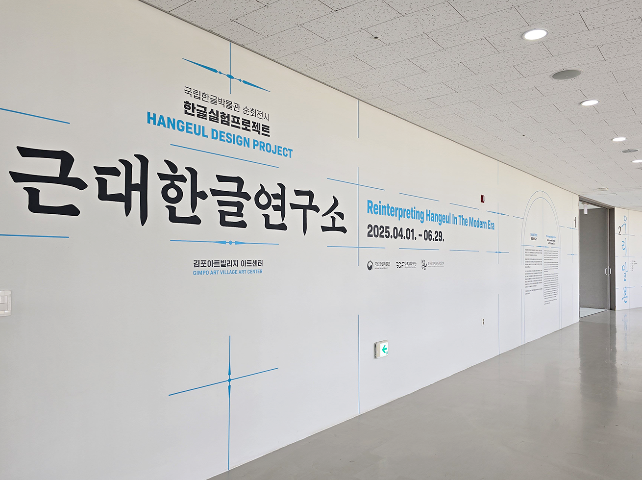 ‘근대한글연구소’ 전시장 입구 사진이다. 하얀색 벽을 따라 긴 복도가 펼쳐져 있다. 벽에는 큼직한 검은색 글씨로 ‘근대한글연구소’라고 쓰여 있다. 