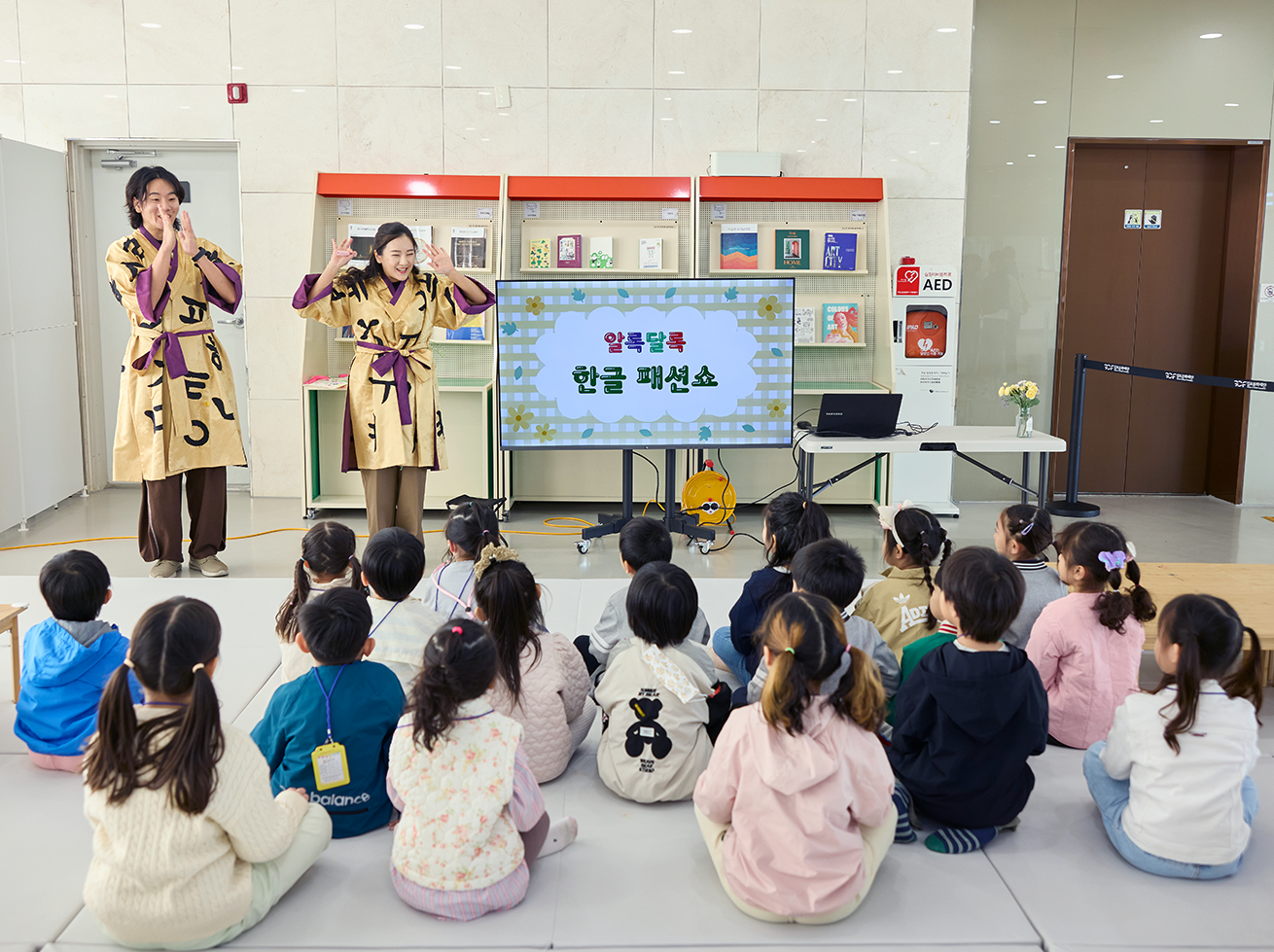 ‘알록달록 한글 패션쇼’ 수업 설명을 듣는 아이들 사진이다. 앞쪽으로 모니터가 있고, 화면에는 꽃무늬 배경에 ‘알록달록 한글 패션쇼’라고 적혀있다. 아이들은 모니터를 바라보고 앉아 있다. 모니터 옆쪽으로는 한글 글자가 큼직하게 인쇄된 전통 의상을 입고 있는 강사 두 명이 아이들을 바라보며 활짝 웃고 있다. 