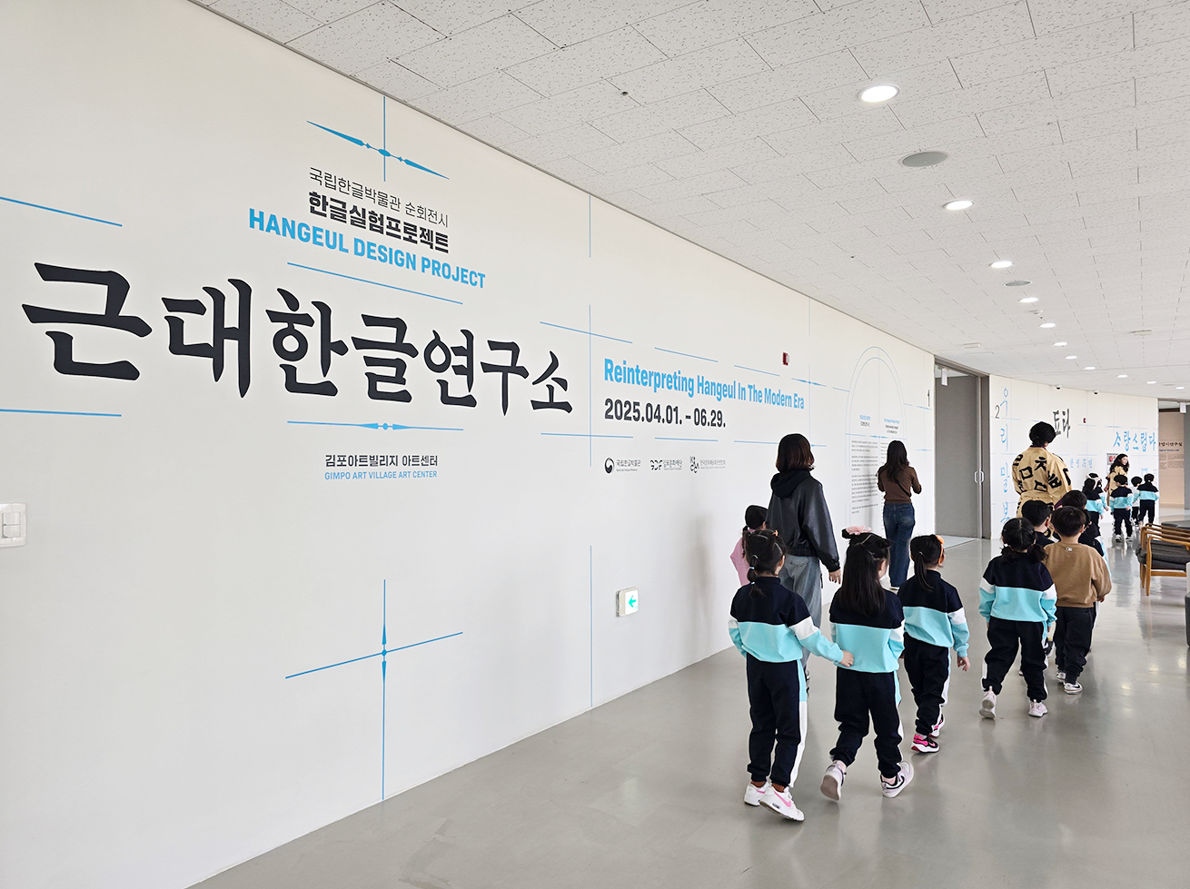 전시실로 이동하는 아이들 사진이다. 아이들은 검은색과 하늘색이 섞인 단체복을 입고 있다. 큼직한 검은색 글씨로 ‘근대한글연구소’라고 적혀있는 벽 앞을 아이들과 선생님들이 걷고 있다.
