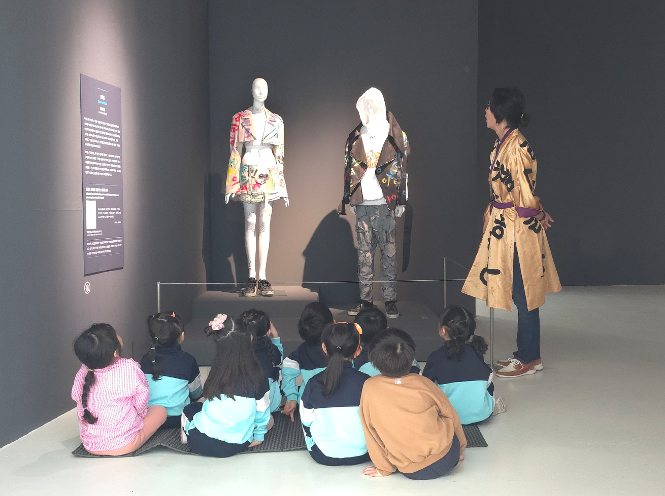 아이들이 작품을 관람하며 설명을 듣는 모습이다. 한글이 프린팅된 화려한 색감의 옷과 검은색 옷을 입은 마네킹 두 개가 전시되어 있다. 아이들은 바닥에 모여 앉아 선생님의 작품 설명을 경청하고 있다.