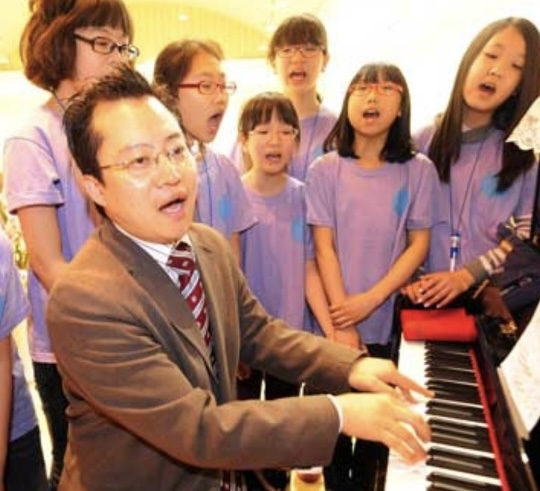 아이들과 함께 노래하고 있는 이강산 작곡가 사진이다. 이강산 작곡가가 피아노 앞에 앉아 연주하며 아이들과 함께 노래를 부르고 있다. 이강산 작곡가 뒤에 서 있는 아이들은 보라색 반팔을 입고 있으며, 열심히 노래를 따라 부르는 중이다.