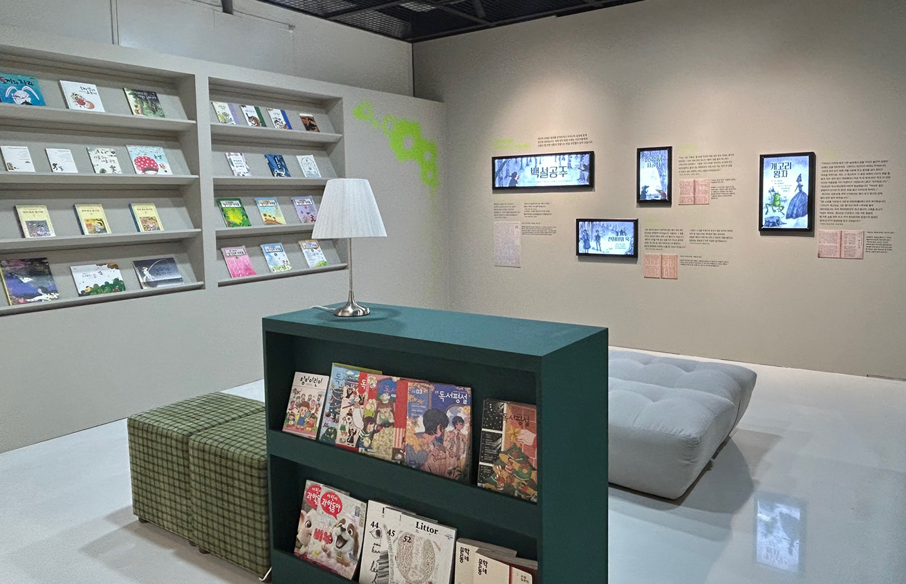 3부 ‘읽고 쓰고 말하는 세상’ 전경 사진이다. 벽면에 책장이 있고, 여러 권의 책이 꽂혀있다. 전시 공간 가운데에는 작은 녹색 책장이 있고, 위에 스탠드 조명이 놓여져 있다. 책을 읽을 수 있는 회색 쇼파와 초록색 체크무늬 의자 두 개가 있다.