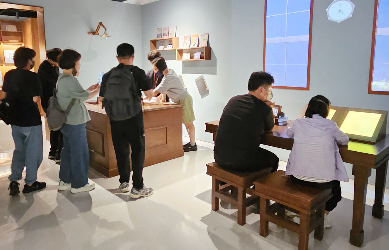 전시에 참여하는 사람들 관람객 사진이다. 왼쪽에 여러 명의 성인과 청소년이 함께 진열장에 전시된 작품을 보고 있고, 오른쪽에는 두 명의 관람객이 나란히 앉아 책상 위 전시 자료를 살펴보고 있다.
