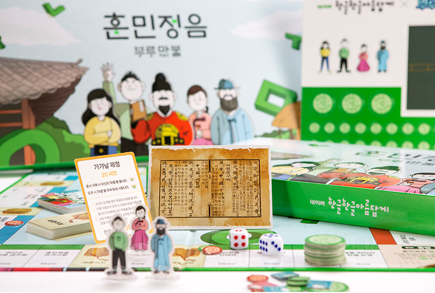 한글 역사 이야기 담은 '부루마불 훈민정음' 특별판 사진이다. 부루마블 형태의 보드판 위에 다양한 게임 소품과 구성품들이 놓여 있다. 뒤쪽 배경에는 훈민정음 부루마블 로고와 세종대왕부터 현대인이 그려진 일러스트가 그려진 박스가 세워져 있다.