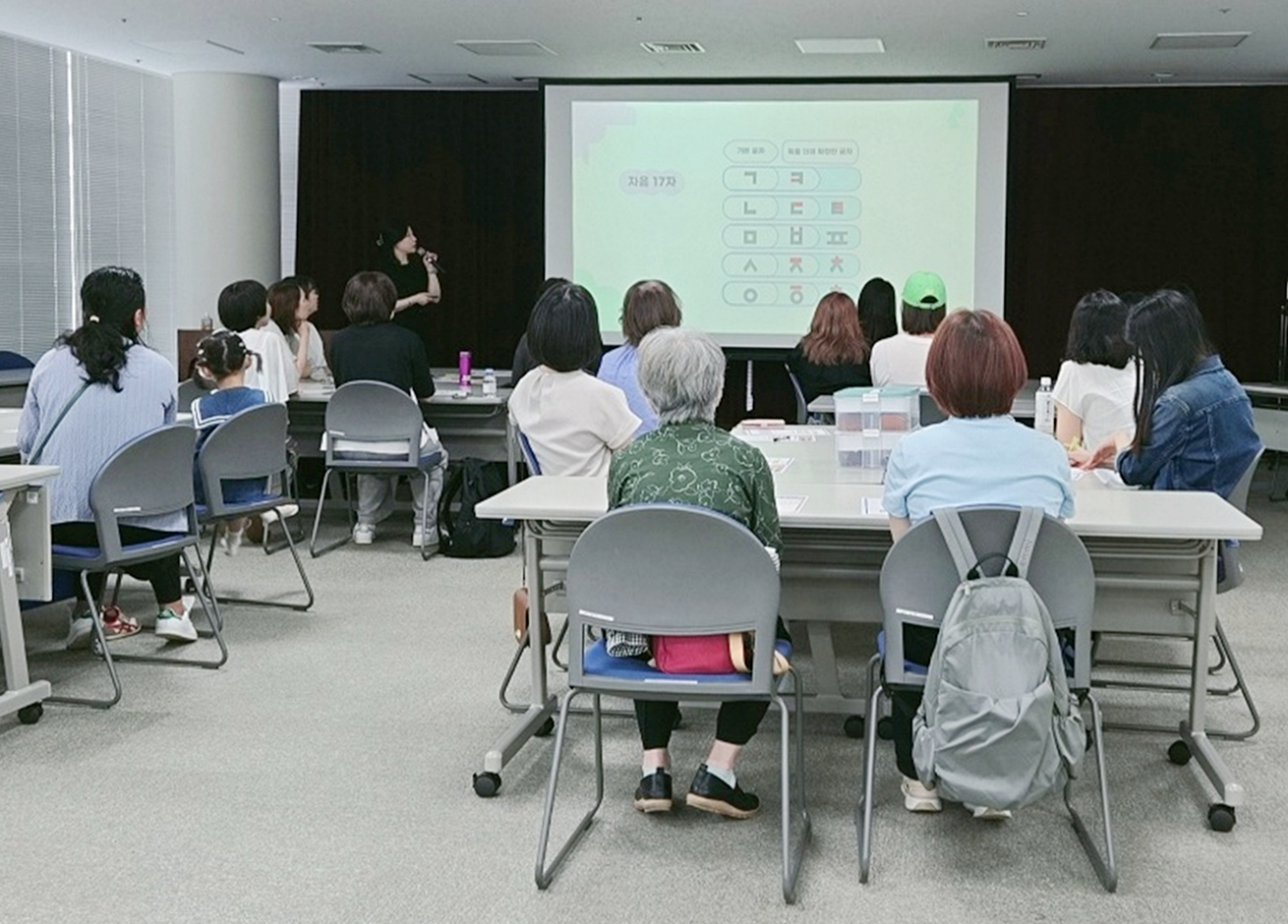 「한글 디자인 공방」 일본 현지 교육 모습 사진이다. 교실 형태의 강의실에서 여러 명의 수강생이 앉아 강의를 듣고 있다. 수강생 대부분은 여성이며, 모두 앞을 바라보고 있고, 각자 앞에 학습 자료와 필기구 등을 두고 있다. 앞쪽 스크린에는 ‘한글 디자인 공방’과 관련된 교육 자료가 빔프로젝터를 통해 출력되어 있으며, 강사가 프레젠테이션을 진행하고 있다.