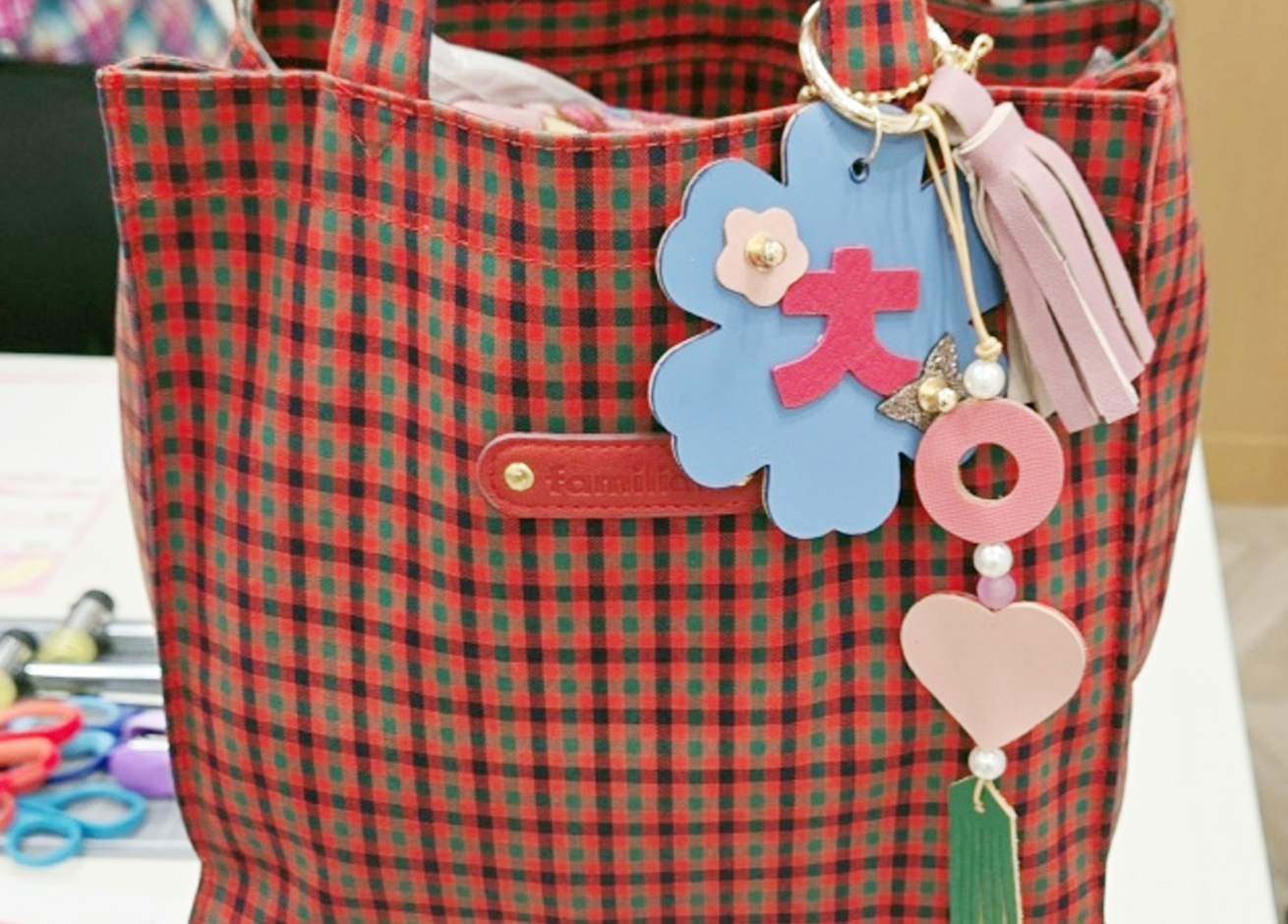 「한글 디자인 공방」 일본 현지 교육 참가자의 작품 사진이다. 체크무늬 가방 손잡이에 한글 키링이 달려 있는 모습이다. 키링에는 자음 'ㅊ', ‘ㅇ’과 꽃, 하트, 태슬이 달려있으며 핑크, 하늘색, 초록색 등이 조화를 이룬다.