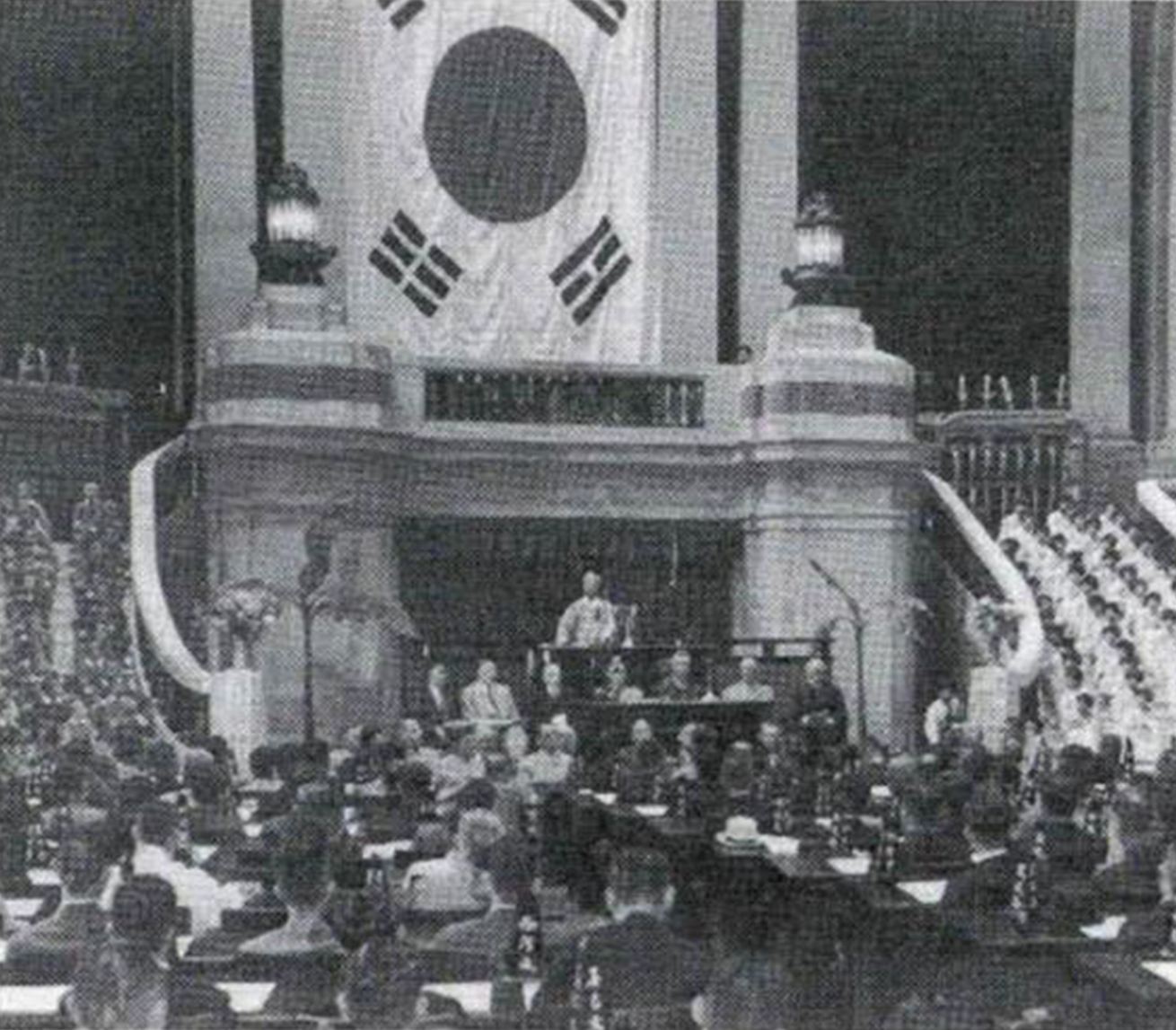 1948년 제헌 국회 개원식 사진이다. 흑백 사진이며, 건물 내부 중앙 무대 위에 커다란 태극기가 걸려 있다. 중앙 단상에는 연설 중인 인물이 서 있고, 그 주변에 내빈들과 국회의원들이 자리를 지키고 있다.