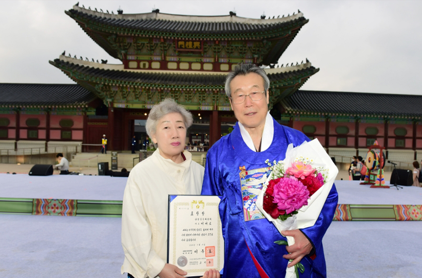 제44회 세종문화상 대통령 표창 수상한 이대로 대표 사진이다. 이대로 대표는 푸른색의 전통 한복을 갖춰 입고 있으며, 손에 꽃다발을 들고 있다. 그 옆에는 흰색 계열의 단정한 옷을 입은 부인이 표창장을 들고 서있다.
