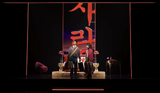 중앙 무대에는 두 인물이 서로 마주 보고 서 있으며, 무대 뒤에는 ‘사랑’이라는 붉은 색의 한글 글씨가 커다랗게 세로로 적혀 있다