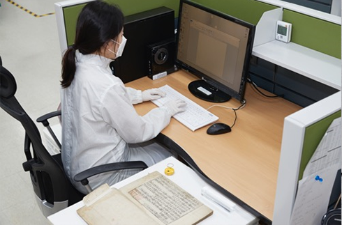 유물 분석 후 입력 작업 사진이다. 작업자가 책상 앞에 앉아 컴퓨터로 유물 자료를 입력하고 있다.