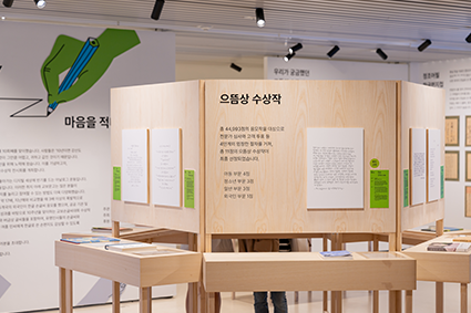 제11회 교보손글씨 대회 체험공간 사진이다. 여러 개의 나무 벽면이 둥글게 배치되어 있고 각 벽면에는 손글씨로 적은 작품들이 걸려 있다. 상단에는 수상작에 대한 간단한 설명이 적혀 있으며, 사람들은 이 공간을 돌아다니며 손글씨 작품을 가까이에서 관람할 수 있다.