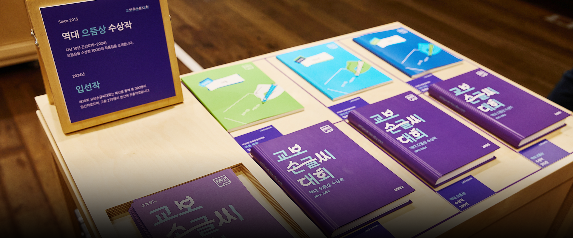 반갑습니다 사진. 책상이 놓인 실내 공간 위에 다양한 색상의 책자들이 가지런히 전시되어 있다. 책자들은 초록색, 하늘색, 파란색, 보라색 등으로 구성되어 있으며, ‘교보 손글씨 대회’ 관련 문구가 표지에 큼직하게 적혀있다. 왼쪽에는 책자들에 대한 설명이 담긴 입간판이 세워져 있고, ‘역대 으뜸상 수상작’이라는 문구가 적혀있다. 왼쪽 하단에는 주황색 박스 안에 ‘반갑습니다’ 문구가 적혀있다. 그 아래에는 기사의 제목 ‘한글과 책으로, 사람과 사람을 잇다 교보문고’가 쓰여있다.