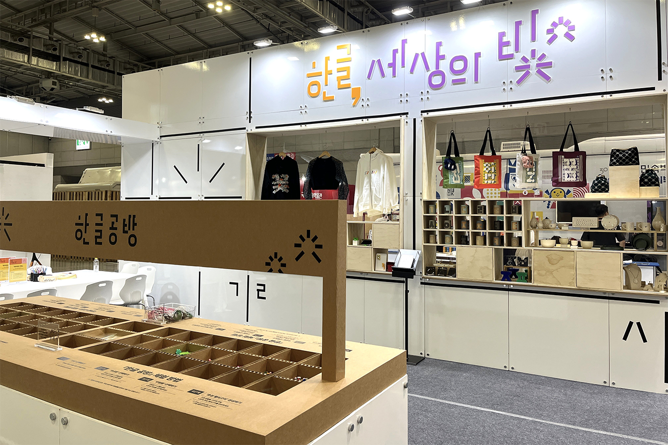 ‘한글, 세상의 빛’ 체험터 현장 사진이다. 부스 상단에는 ‘한글, 세상의 빛’이라는 문구가 주황색과 보라색으로 디자인되어 크게 걸려있다. 내부에는 다양한 전시 및 체험 요소가 배치되어 있는데, 앞쪽에는 ‘한글공방’이라고 적혀있는 여러 칸으로 나뉜 갈색 구조물이 놓여 있다. 뒤쪽 벽에는 티셔츠와 에코백 같은 상품들이 전시되어 있고, 칸칸이 나뉜 진열장에는 소품과 기념품들이 가지런히 놓여 있다.