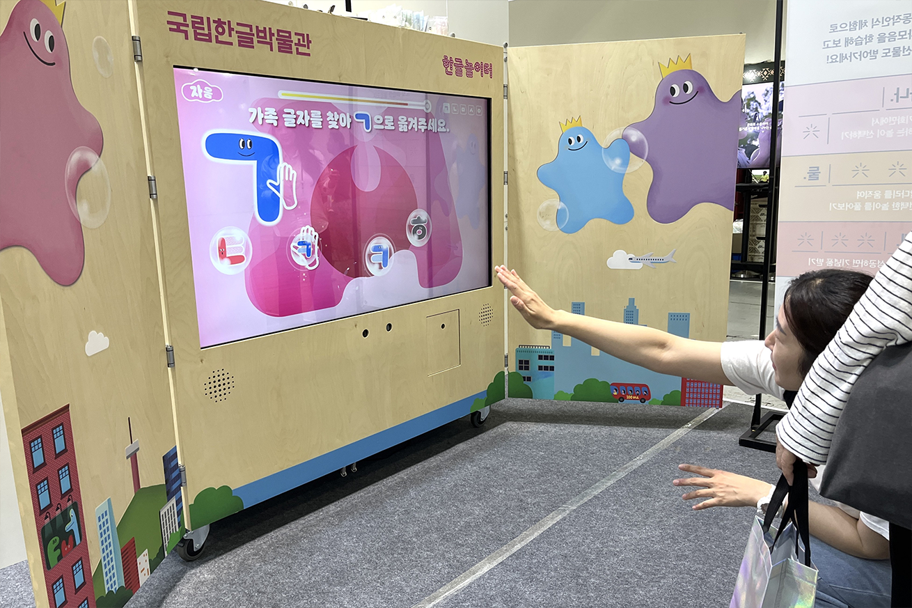 ‘안녕! 한글 놀이터’ 현장 사진이다. 중앙에는 스크린이 설치되어 있으며, 화면에는 분홍색과 파란색 캐릭터가 등장하고 ‘가족 글자를 찾아 ㄱ으로 옮겨주세요’라는 안내 문구가 보인다. 화면 앞에서는 한 여성이 오른손을 뻗어 동작 인식을 통해 게임에 참여하고 있다.