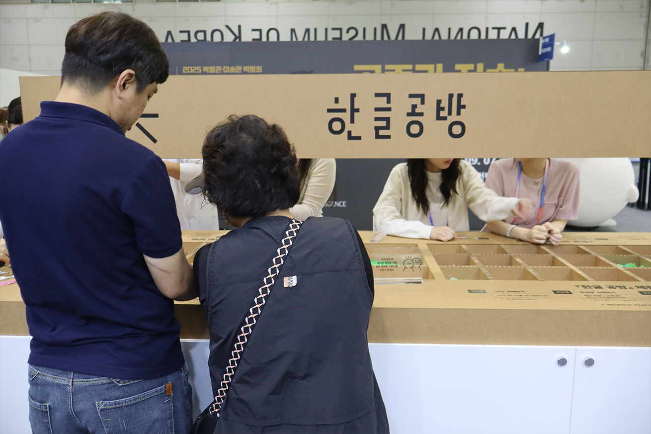 ‘한글공방’ 현장 사진이다. 갈색 종이 재질의 큰 칸막이에 굵은 글씨로 ‘한글공방’이라고 쓰여 있다. 자음과 다양한 모양의 구슬이 칸막이 안에 들어가 있다. 앞쪽에는 남성과 여성이 나란히 서서 체험 칸을 들여다보고 있다.