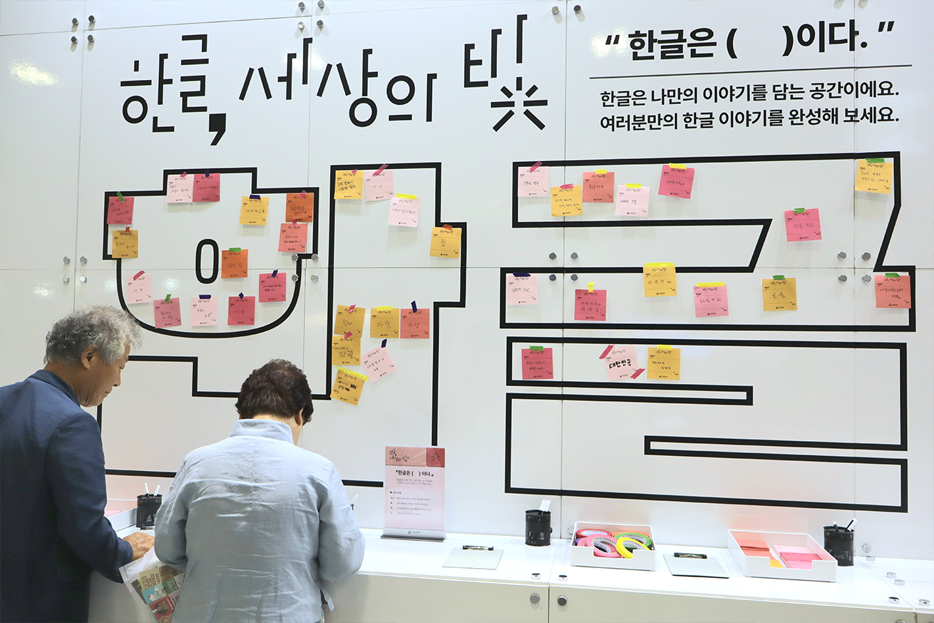‘한글은 세상의 ( )이다’ 현장 사진이다. 흰 벽면에 커다란 검은색 글씨로 ‘한글, 세상의 빛’이라는 문구와 함께 ‘한글은 ( )이다.’라는 문장이 적혀있다. 벽면 중앙에는 큼지막하게 ‘한글’ 글자가 디자인되어 있고, 그 위에 분홍색·주황색·노란색 메모지가 붙어 있다. 두 사람이 탁자 앞에 서서 메모지에 글을 적고 있다.