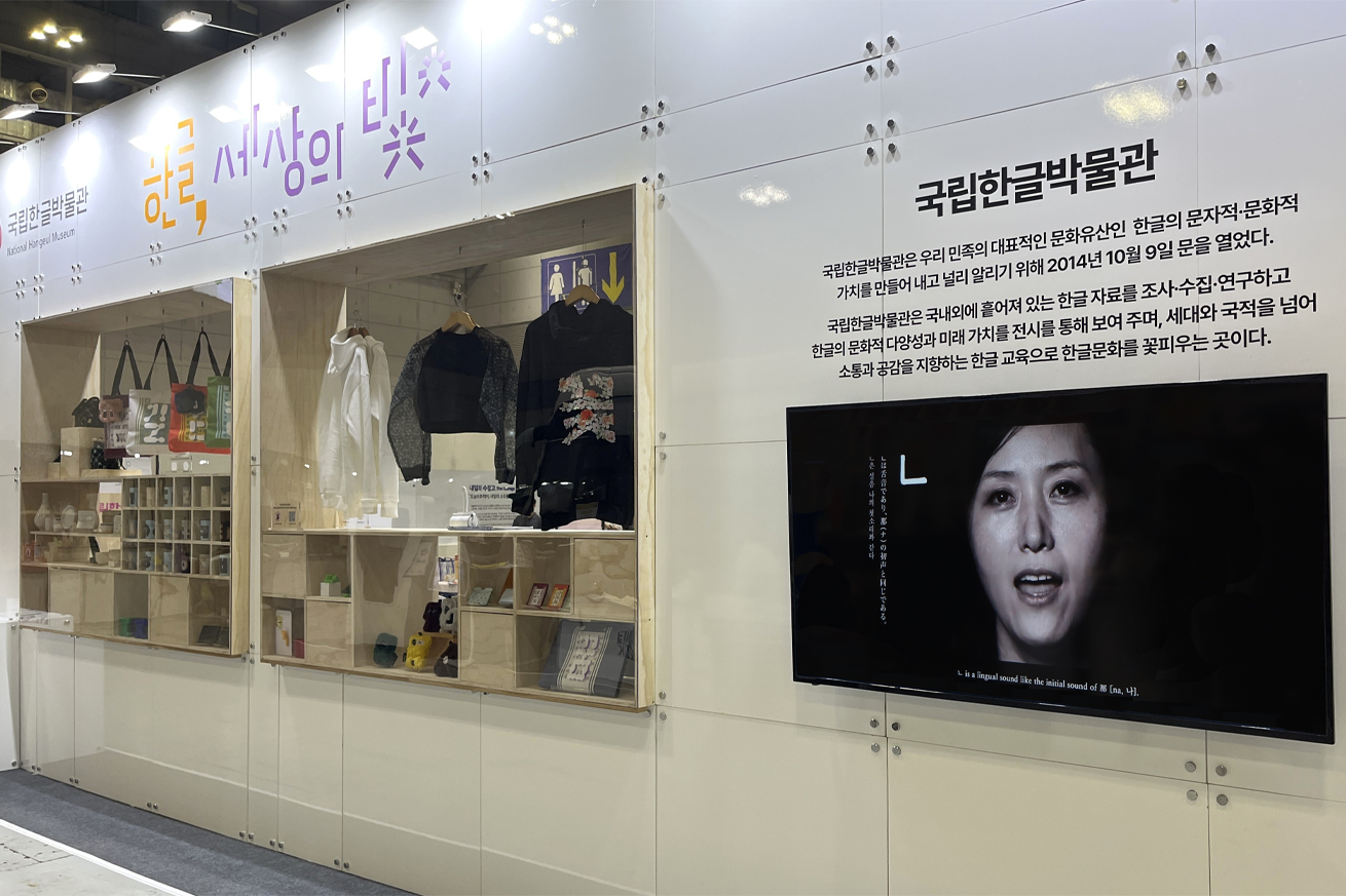 한글 문화상품 전시 사진이다. 벽 상단에는 보라색과 주황색 글씨로 ‘한글, 세상의 빛’이라는 큰 문구가 적혀있다. 그 아래에는 투명한 진열장이 설치되어 다양한 전시품이 전시되어 있다. 진열장 안에는 한글 관련 기념품, 책, 옷, 가방 등이 가지런히 놓여 있다. 그 오른쪽으로는 한글 체험을 설명하는 TV가 걸려있다. 화면 속에는 ‘니은’을 발음하는 여성의 얼굴이 크게 비춰지고 있다.