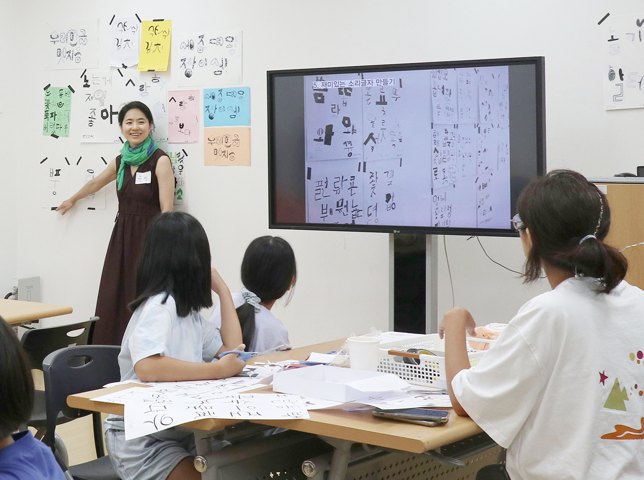 ‘우리 한글 멋지음’ 교육 현장 사진이다. 교사가 모니터 화면과 벽에 붙은 글자 자료를 활용해 아이들에게 설명하고 있고, 학생들은 집중해 듣고 있다.