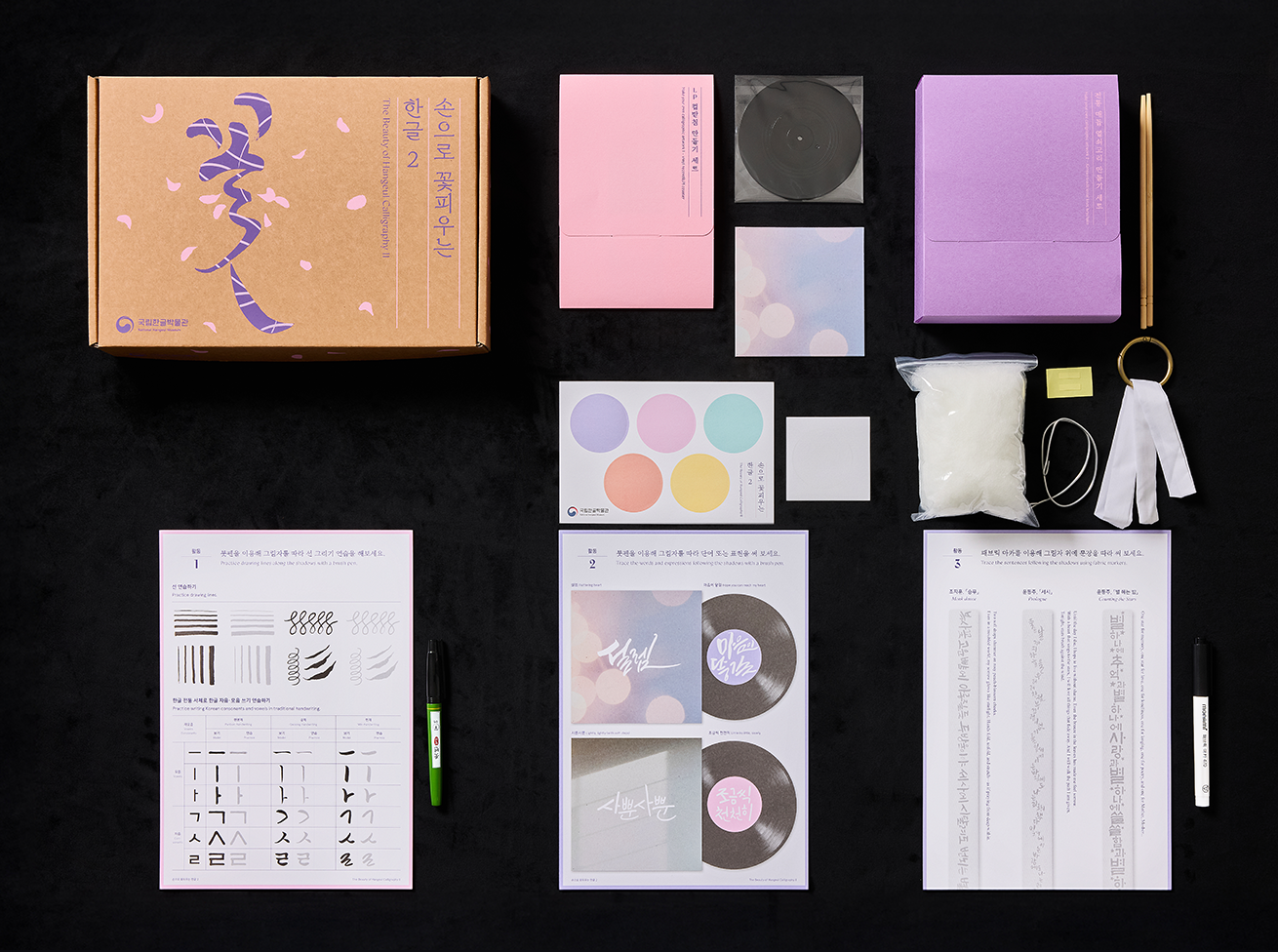 ‘손으로 꽃피우는 한글 2 (The Beauty of Hangeul CalligraphyⅡ)>’ 교육 사진이다. 보라색과 분홍색 종이, 붓, 잉크, 안내문 등 다양한 재료가 정갈히 놓인 키트가 놓여 있다.
