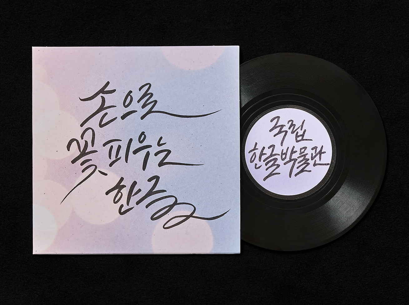 ‘손으로 꽃피우는 한글 2 (The Beauty of Hangeul CalligraphyⅡ)>’ 교육 사진이다. LP판 모양의 원반과 함께 ‘손으로 꽃피우는 한글’이라는 캘리그래피가 쓰인 종이가 놓여 있다.